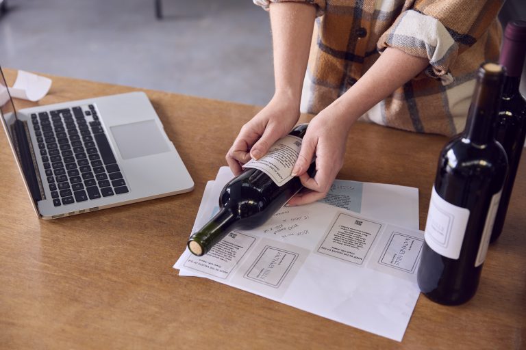 Persoană aplicând o etichetă cu cod QR pe o sticlă de vin, lângă un laptop și alte sticle etichetate.