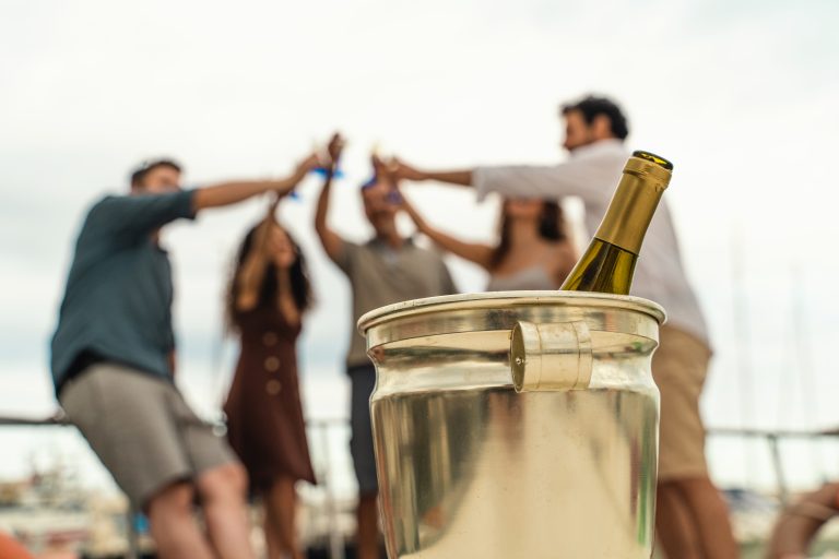 Sticlă de vin alb într-un recipient cu gheață, în prim-plan, cu un grup de prieteni tineri ciocnind în fundal