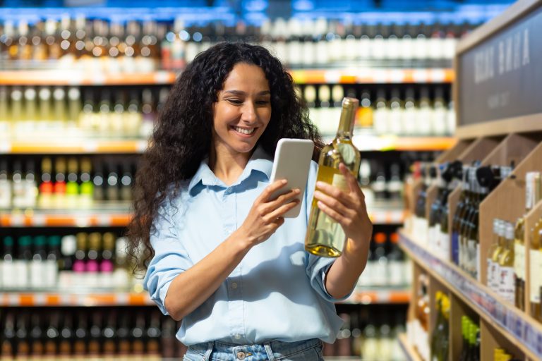 Femeie zâmbitoare scanează cu telefonul eticheta unei sticle de vin într-un supermarket.