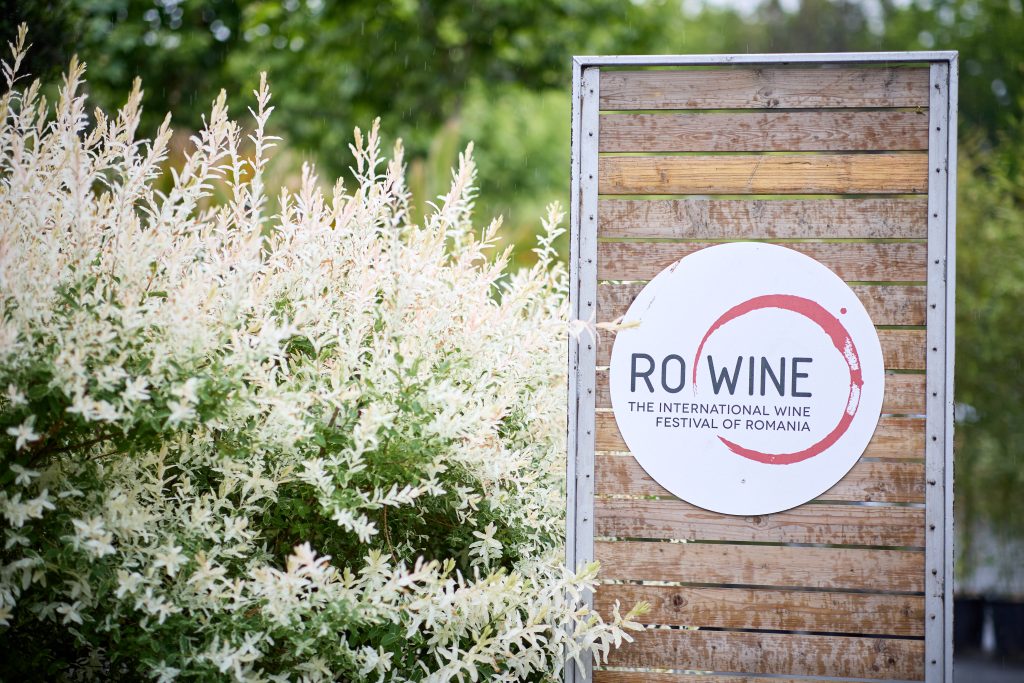 Panou cu sigla RO-Wine – The International Wine Festival of Romania, amplasat în zona de intrare, înconjurat de vegetație